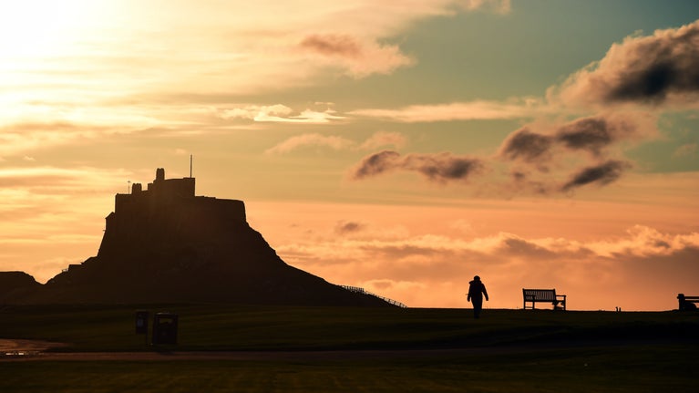 A sunrise photograph of Lindisfarne from Holy Island harbour with a silhouetted figure and a bench in the foreground